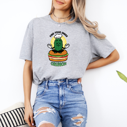 Person wearing a gray t-shirt with a graphic design of a pickle on a hamburger, text 'Find your inner crunch', and blue jeans.